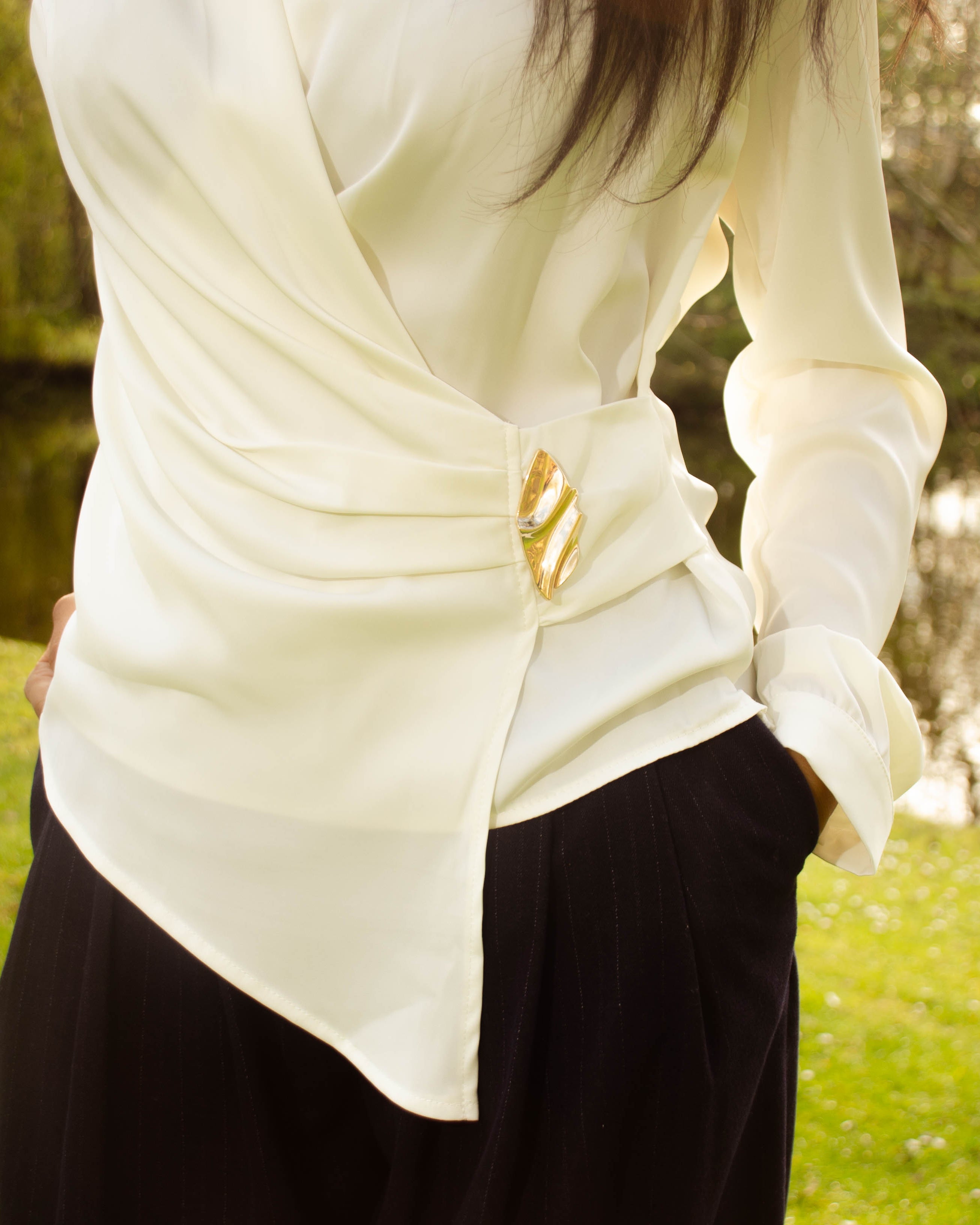 A person outdoors by a lake wearing a cream wrap-style blouse with a gold decorative accent at the waist.